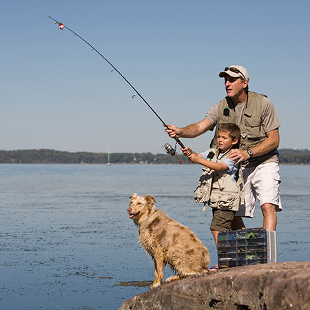 Father teaching fly fishing with son on the rocks with family dog nearby.